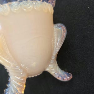 May include: A pale, translucent opalescent glass vessel featuring a smooth, rounded body. The rim is decorated with a series of raised, circular beads, and the base is supported by three curved, textured legs with a distinct dotted pattern.