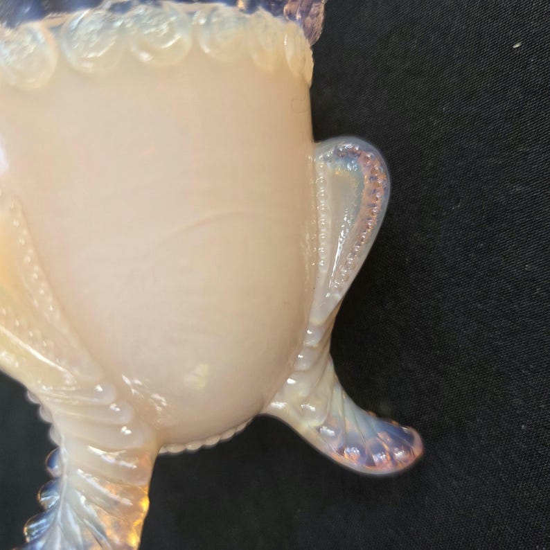 May include: A pale, translucent opalescent glass vessel featuring a smooth, rounded body. The rim is decorated with a series of raised, circular beads, and the base is supported by three curved, textured legs with a distinct dotted pattern.