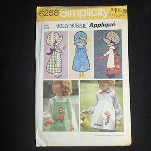May include: A Simplicity 6258 sewing pattern envelope for Holly Hobbie appliqués. The cover features illustrations of three characters in bonnets and dresses, alongside photographs of children wearing garments decorated with these appliqués.