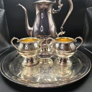 May include: A silver-plated tea service set comprising a tall teapot, a creamer, and a sugar bowl, all arranged on a round, ornate silver-plated serving tray. Each piece features a polished metallic finish with gold-toned interior accents.