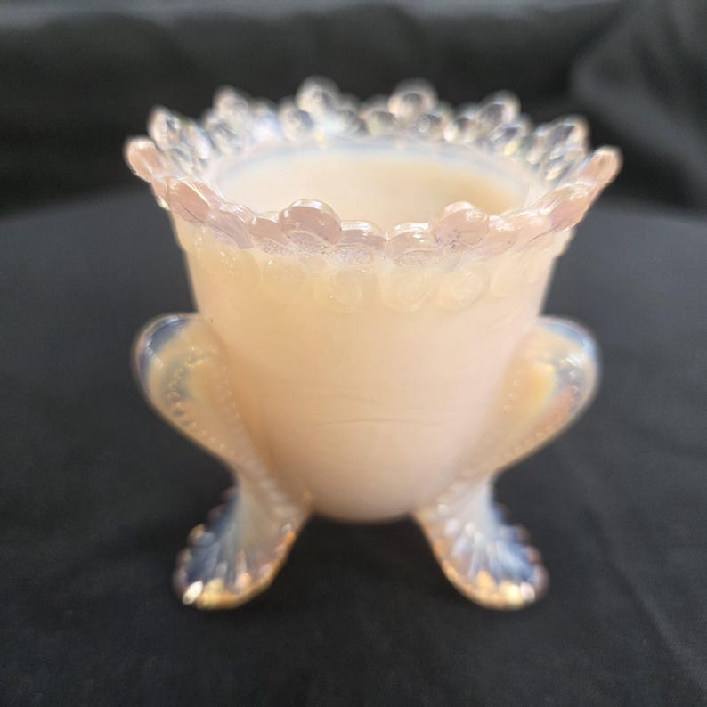 May include: A small, translucent opalescent glass vessel with a soft peach hue. The piece features a scalloped, beaded rim and stands on three curved, textured feet that extend upward along the sides of the body, creating a distinct tripod base.
