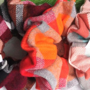 May include: A collection of plaid scrunchies in various colors, including red, orange, gray, and teal. Each scrunchie features a unique plaid pattern and a small white tag with the text "Scrunchy Sun Shop".