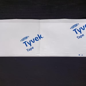 May include: White Tyvek tape with blue "DuPont Tyvek Tape" text. The tape is folded in half and lies on a black surface. The tape is used for construction and packaging.