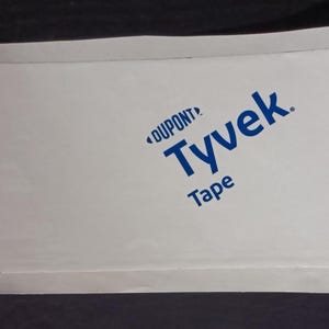 May include: White rectangular roll of Tyvek tape with blue text that reads "DUPONT Tyvek Tape". The tape is folded over itself. The background is dark.