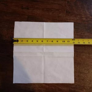 Puede incluir: Un trozo de papel blanco cuadrado doblado por la mitad, medido con una cinta métrica amarilla. La cinta métrica indica 30,5 cm.