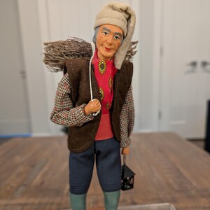 May include: A handcrafted figurine of a man carrying a bundle of twigs on his back and a lantern. He wears a red shirt, brown vest, and blue pants. The figure has a beige hat and green socks. The figurine is standing on a wooden base.