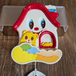 May include: A colorful baby mobile featuring a house-shaped design with a smiling face, red roof, and blue eyes. The mobile includes yellow teddy bear figures and a cloud-shaped accent. A white handle hangs below.