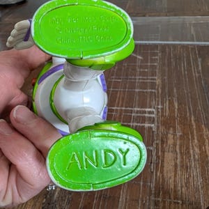 May include: A plastic Buzz Lightyear action figure with green feet. One foot is embossed with the name ANDY in capital letters. The other foot features molded text reading Mfg. For McD Corp. &copy; Disney/Pixar China TRC Chine. The figure is white and purple.