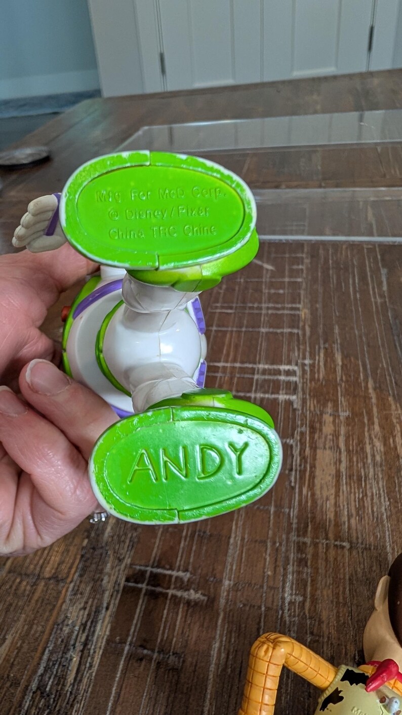 May include: A plastic Buzz Lightyear action figure with green feet. One foot is embossed with the name ANDY in capital letters. The other foot features molded text reading Mfg. For McD Corp. &copy; Disney/Pixar China TRC Chine. The figure is white and purple.