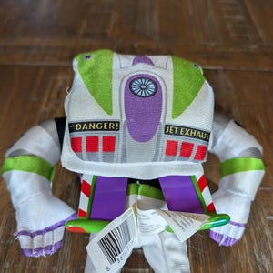 May include: A plush Buzz Lightyear toy, featuring a white and green spacesuit design. The toy has purple accents, the words "DANGER!" and "JET EXHAUS" printed on it, and a barcode tag.