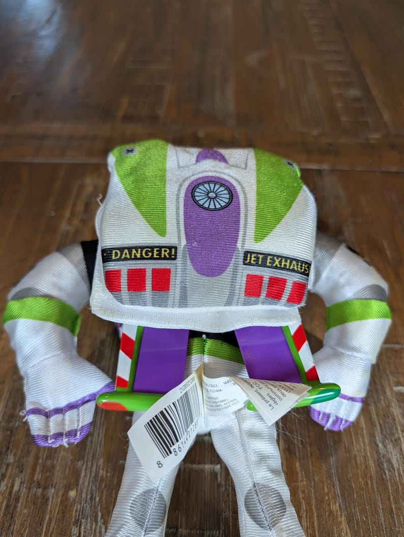 May include: A plush Buzz Lightyear toy, featuring a white and green spacesuit design. The toy has purple accents, the words "DANGER!" and "JET EXHAUS" printed on it, and a barcode tag.