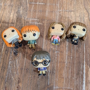 May include: Five vinyl figurines of characters from the Harry Potter series. The figures have oversized heads and small bodies. The characters include Hermione, Ron, Harry, and two other female characters, all with distinctive hairstyles and outfits. They are arranged on a wooden surface.