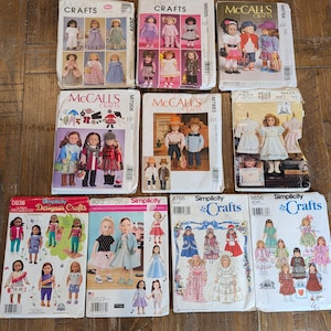 May include: A collection of eleven sewing pattern envelopes for doll clothing, featuring brands McCall's Crafts, Simplicity, and Vogue. Each rectangular paper envelope displays color illustrations of various doll outfits, including dresses, hats, and casual wear, with printed brand names and pattern numbers.