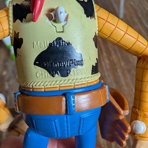 May include: A plastic cowboy action figure with a yellow shirt featuring black cow spots, a red neckerchief, and blue pants. The back of the shirt displays molded text reading Mattel, Inc. Disney/Pixar CHINA 1996, with a pull-string ring at the top.