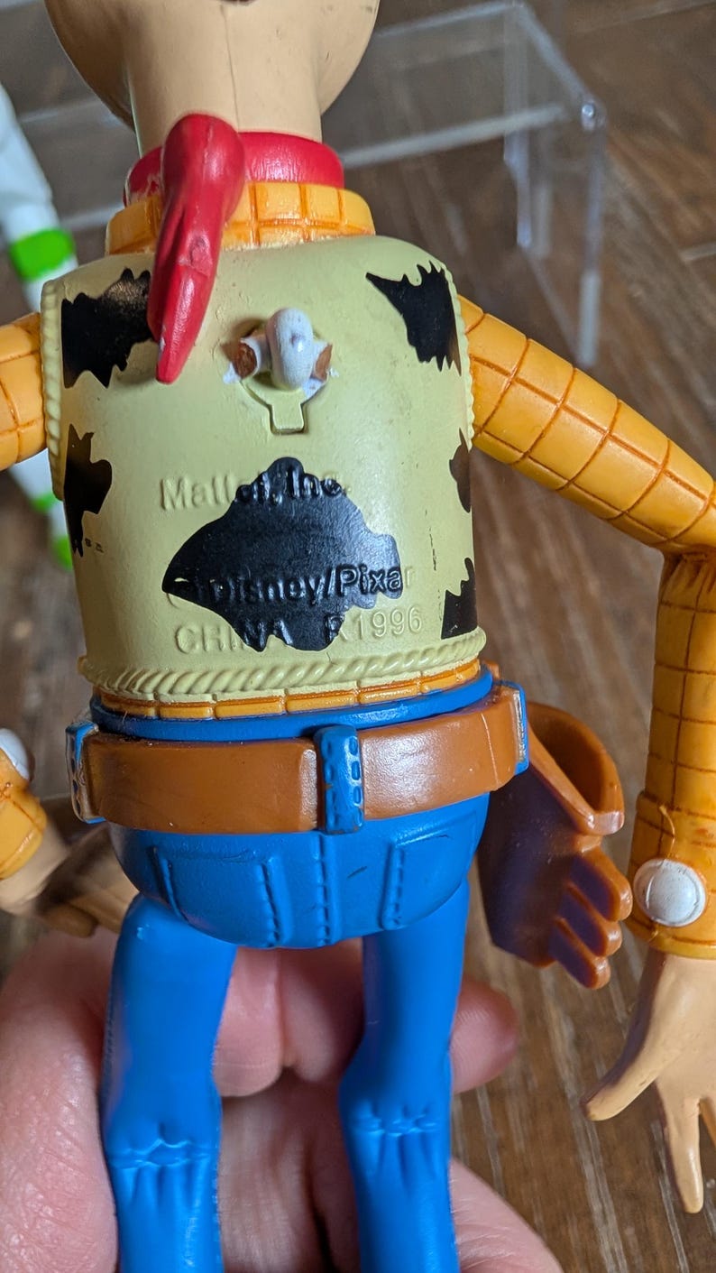 May include: A plastic cowboy action figure with a yellow shirt featuring black cow spots, a red neckerchief, and blue pants. The back of the shirt displays molded text reading Mattel, Inc. Disney/Pixar CHINA 1996, with a pull-string ring at the top.