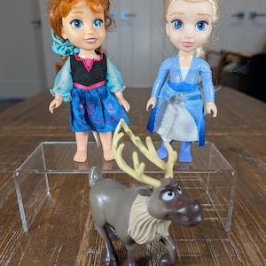 May include: Three toy figures on a wooden surface. Two dolls, one with red hair and a blue dress, the other with blonde hair and a blue dress. A brown reindeer toy with large antlers is in front.