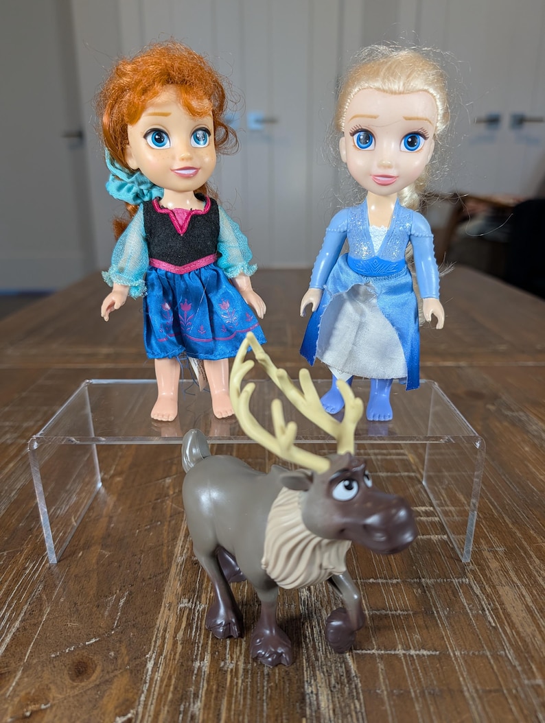 May include: Three toy figures on a wooden surface. Two dolls, one with red hair and a blue dress, the other with blonde hair and a blue dress. A brown reindeer toy with large antlers is in front.
