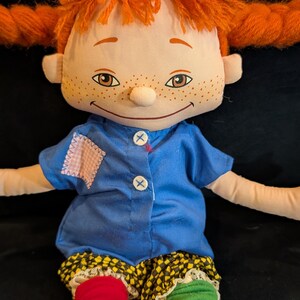 Op de afbeelding: Een handgemaakte lappenpop met oranje garen haar in vlechtjes. De pop heeft een blauw shirt, geel en zwart geruite shorts en mismatched rode en groene sokken. Het gezicht van de pop heeft sproeten en een gestikte glimlach.
