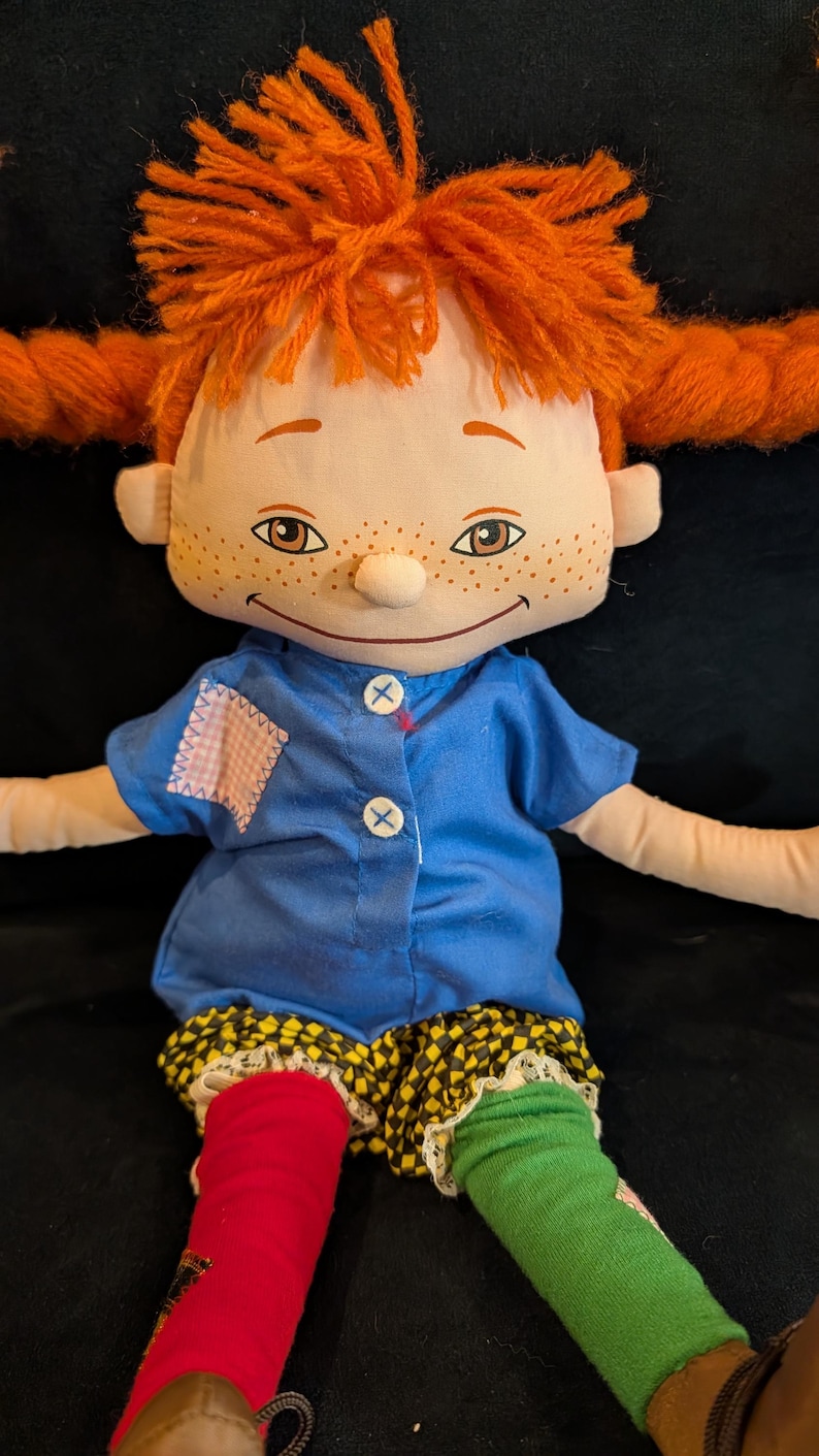 Op de afbeelding: Een handgemaakte lappenpop met oranje garen haar in vlechtjes. De pop heeft een blauw shirt, geel en zwart geruite shorts en mismatched rode en groene sokken. Het gezicht van de pop heeft sproeten en een gestikte glimlach.