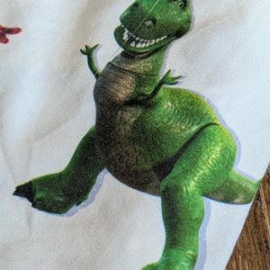 May include: A light blue fabric featuring a repeating pattern of a green Tyrannosaurus rex dinosaur with a wide, toothy grin. The design also includes yellow stars outlined in red and small blue stars scattered across the light blue background.
