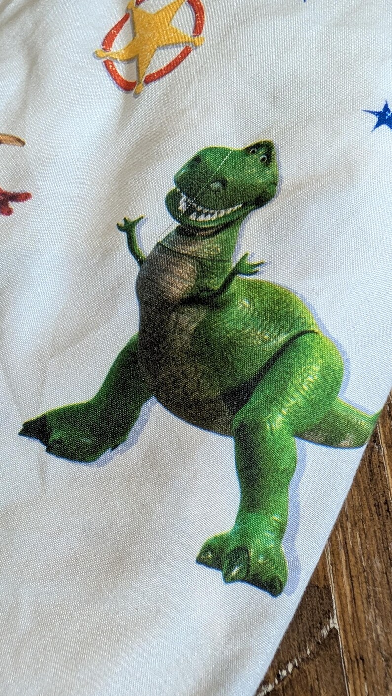 May include: A light blue fabric featuring a repeating pattern of a green Tyrannosaurus rex dinosaur with a wide, toothy grin. The design also includes yellow stars outlined in red and small blue stars scattered across the light blue background.