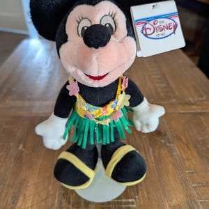 May include: A Minnie Mouse plush toy wearing a Hawaiian-style outfit. The plush has black ears, a pink face, and a red mouth. It is wearing a yellow and green hula skirt and a lei. A tag from The Disney Store is attached.