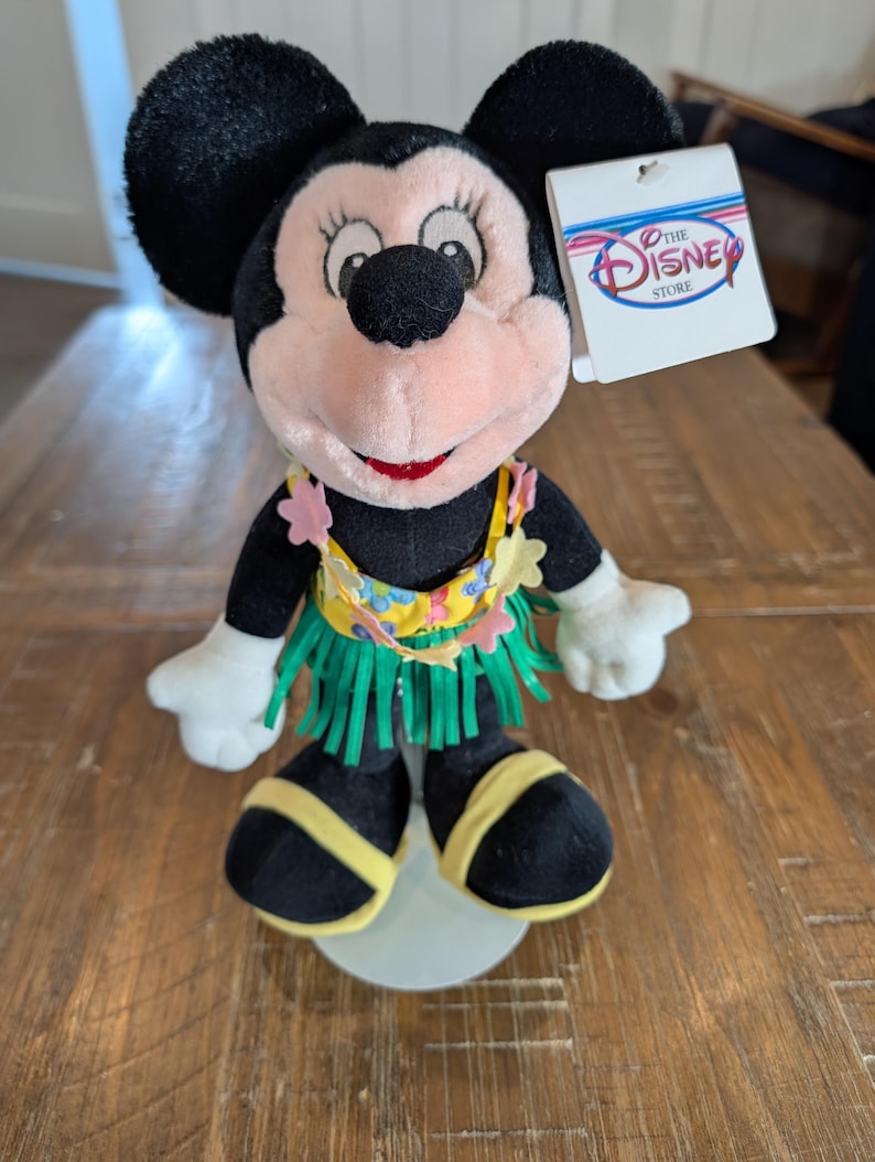 May include: A Minnie Mouse plush toy wearing a Hawaiian-style outfit. The plush has black ears, a pink face, and a red mouth. It is wearing a yellow and green hula skirt and a lei. A tag from The Disney Store is attached.