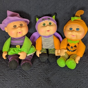 May include: Three dolls dressed in Halloween costumes. The first wears a purple witch hat and green skirt. The second is in a purple and orange bat costume with green ears. The third wears an orange pumpkin hat and a pumpkin-faced orange jumpsuit.
