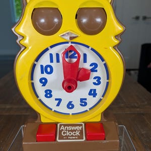 May include: A yellow and brown owl-shaped plastic learning clock with a white face, blue numbers, and red hands. The base features two red buttons and a white label that reads Answer Clock by TOMY. The clock has two large brown eyes.