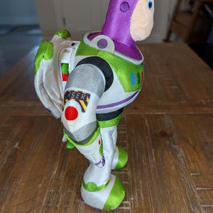 May include: A Buzz Lightyear toy figure, featuring a white and green spacesuit with a purple hood and a smiling face. The figure has a "LASER" label on the arm and a pair of white wings.