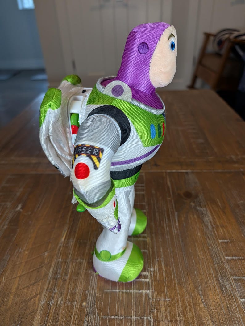 May include: A Buzz Lightyear toy figure, featuring a white and green spacesuit with a purple hood and a smiling face. The figure has a "LASER" label on the arm and a pair of white wings.