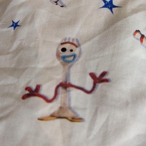 May include: A white fabric featuring a repeating pattern of Toy Story characters including Forky, a white spork figure with red pipe cleaner arms, a blue mouth, and mismatched eyes, alongside Buzz Lightyear, yellow planets, and blue stars.