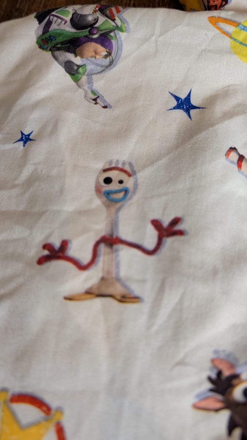 May include: A white fabric featuring a repeating pattern of Toy Story characters including Forky, a white spork figure with red pipe cleaner arms, a blue mouth, and mismatched eyes, alongside Buzz Lightyear, yellow planets, and blue stars.