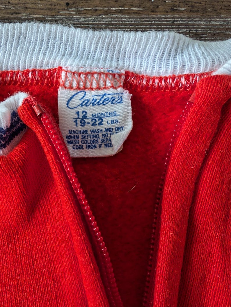 May include: Red Carter's brand zippered sleeper with white trim and a white tag that reads "Carter's 12 Months 19-22 LBS Machine Wash and Dry Warm Setting, No Wash Colors Separately Cool Iron If Needed".