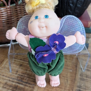 May include: A vintage doll with a round face, blue eyes, and blonde hair styled in a bun. The doll wears a pink top, green velvet pants, and purple flower accents. It has iridescent wings and is standing on a clear display stand.