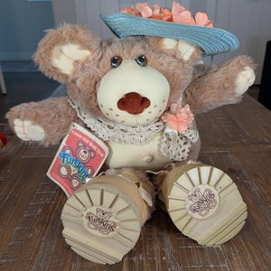May include: A plush teddy bear with light brown fur, a cream-colored muzzle, and a dark brown nose. It wears a light blue straw hat with peach-colored flowers, a cream lace collar, and tan boots with Furskins branding on the soles. A Furskins tag is attached.