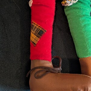 Op de afbeelding: Close-up van de benen van een pop. E&eacute;n been is rood met een geruite patch, de andere is groen. De pop draagt bruine schoenen met bruine veters. De kleding van de pop bevat geel en zwart geruite stof.