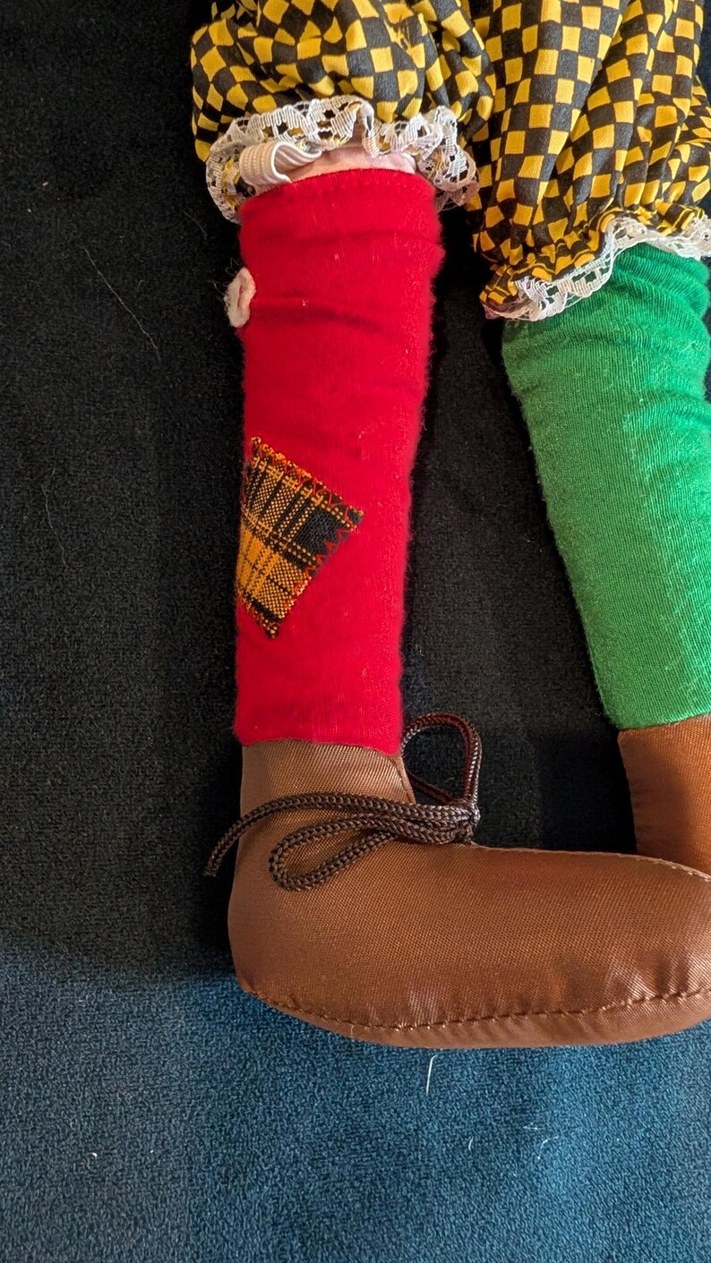 Op de afbeelding: Close-up van de benen van een pop. E&eacute;n been is rood met een geruite patch, de andere is groen. De pop draagt bruine schoenen met bruine veters. De kleding van de pop bevat geel en zwart geruite stof.