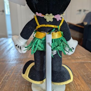 May include: A plush Minnie Mouse doll, viewed from the back, wearing a Hawaiian-themed outfit. The doll has a black body, white gloves, and black shoes with yellow trim. The outfit includes a green grass skirt, a yellow top, and a flower lei.