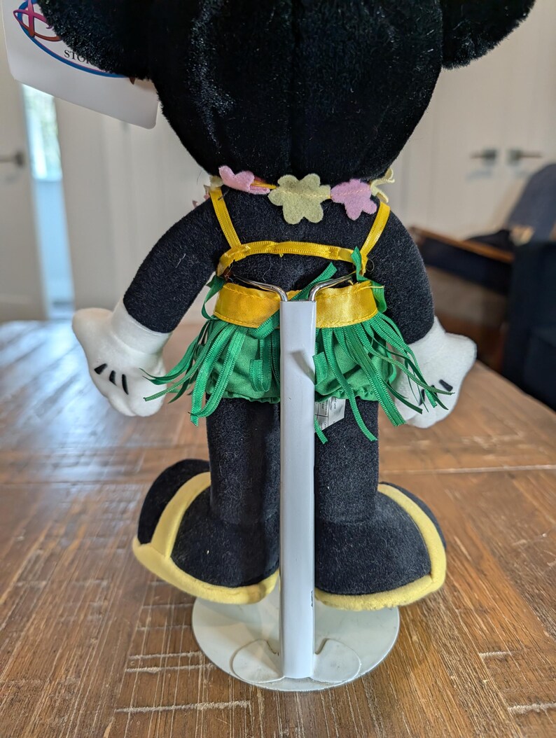 May include: A plush Minnie Mouse doll, viewed from the back, wearing a Hawaiian-themed outfit. The doll has a black body, white gloves, and black shoes with yellow trim. The outfit includes a green grass skirt, a yellow top, and a flower lei.
