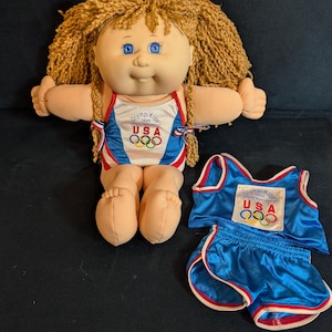 May include: A vintage Cabbage Patch Kids doll with blonde yarn hair and blue eyes, wearing a USA Olympic Kids 1996 outfit. The outfit includes a blue and white tank top and matching shorts.