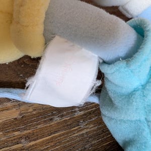 Op de afbeelding: Close-up van een pluche speelgoed met lichtblauwe, gele en witte delen. Een wit label met vervaagde roze tekst is aan het speelgoed bevestigd. Het speelgoed is gemaakt van zachte, getextureerde stof.