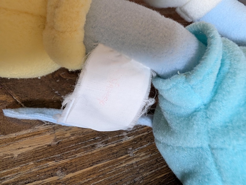 Op de afbeelding: Close-up van een pluche speelgoed met lichtblauwe, gele en witte delen. Een wit label met vervaagde roze tekst is aan het speelgoed bevestigd. Het speelgoed is gemaakt van zachte, getextureerde stof.