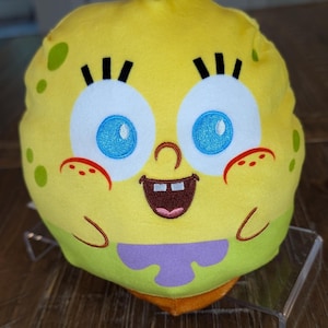 May include: A round, yellow plush toy featuring the embroidered face of SpongeBob SquarePants with large blue eyes, black eyelashes, a brown mouth with two white teeth, and red freckles. The plush has green spots and a purple tie at the bottom.