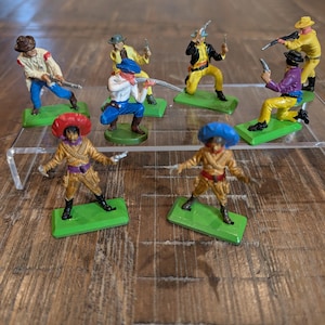 May include: A collection of vintage toy cowboy figures. The figures are painted in various colors, including yellow, blue, and red, and are posed in action stances. Each figure is mounted on a green base, and some hold guns.