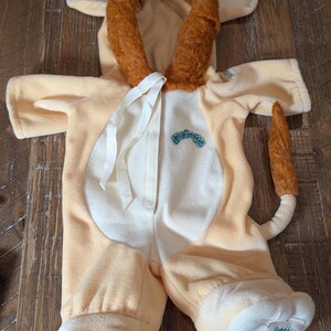May include: A light yellow plush onesie featuring a hood with a brown faux fur lion mane, white ribbon ties, and a matching brown faux fur tail. The garment displays the Cabbage Patch Kids logo in green embroidery on the chest and on the foot.