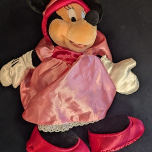 Peut inclure: Poup&eacute;e en peluche Minnie Mouse v&ecirc;tue d'une robe en satin rose et d'un bonnet assorti. La poup&eacute;e a des oreilles noires, un nez noir et des chaussures rouges. Les gants de la poup&eacute;e sont blancs.