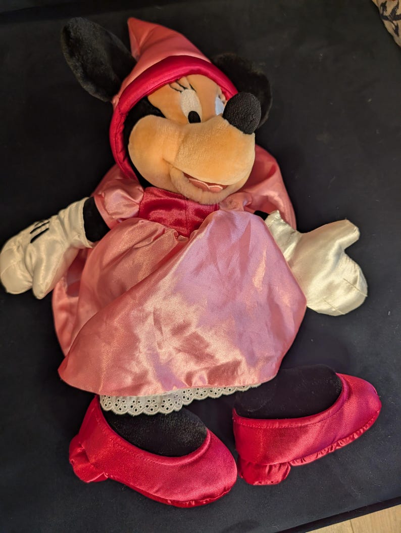 Peut inclure: Poup&eacute;e en peluche Minnie Mouse v&ecirc;tue d'une robe en satin rose et d'un bonnet assorti. La poup&eacute;e a des oreilles noires, un nez noir et des chaussures rouges. Les gants de la poup&eacute;e sont blancs.