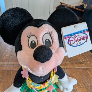 May include: A Minnie Mouse plush toy with black ears, a pink face, and a yellow and green grass skirt. The plushie has a floral lei and a Disney Store tag.
