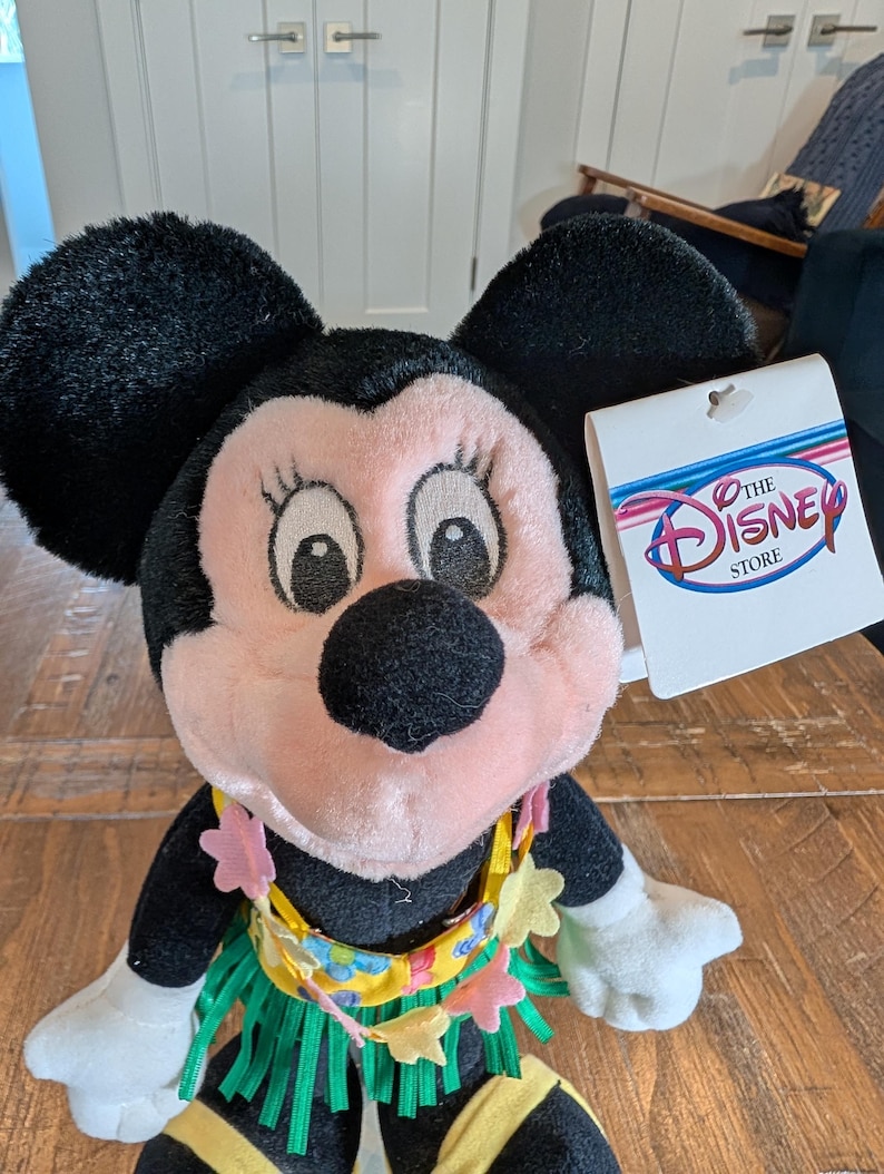 May include: A Minnie Mouse plush toy with black ears, a pink face, and a yellow and green grass skirt. The plushie has a floral lei and a Disney Store tag.
